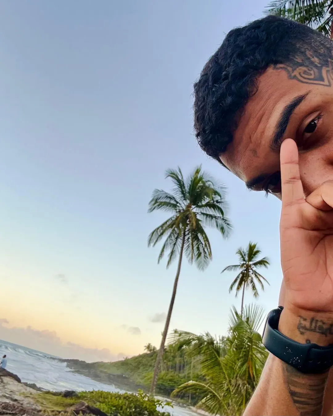 Erick com a mão no rosto, olhando para a câmera, com coqueiros e o mar ao fundo no fim de tarde
